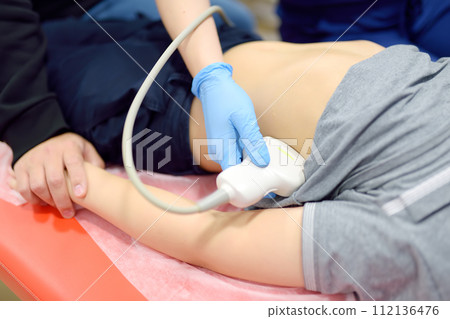 Doctor making abdominal ultrasound for boy using scanner machine. Female doc runs ultrasound sensor over patient tummy. Mother supports son during diagnosis examination of internal organs 112136476