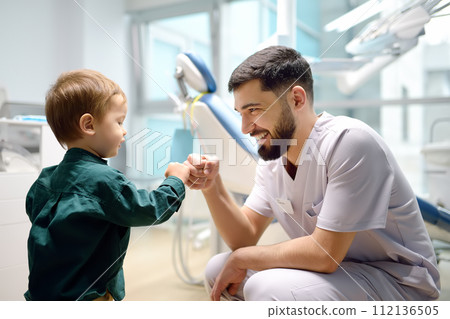 Friendly dentist greets cute toddler boy in office. Doctor encouraging with small child patient and giving him "high five". Kids doc establishes contact with little patient Friendly dentist greets cute toddler boy in office. Doctor encouraging with small child patient and giving him "high five". Kids doc establishes contact with little patient 112136505