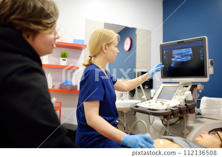 Doctor making abdominal ultrasound for boy using scanner machine. Doc runs ultrasound sensor over patient tummy looking at screen. Mother supports son during examination of internal organs 112136508