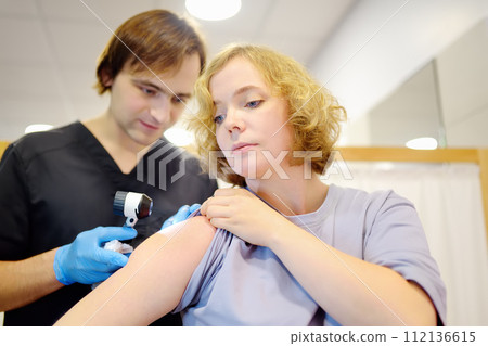 A caring doctor checks moles on the skin of a young woman. A dermatologist looks at a rash on the shoulder of using a dermatoscope. Prevention and diagnosis of melanoma, skin cancer. A caring doctor checks moles on the skin of a young woman. A dermatologist looks at a rash on the shoulder of using a dermatoscope. Prevention and diagnosis of melanoma, skin cancer. 112136615