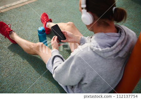 Overhead view of active sportsman wearing headphones, checking sports app on his smart mobile phone Overhead view of active sportsman wearing headphones, checking sports app on his smart mobile phone 112137754