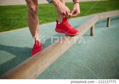 Cropped view of a fit man tying shoelaces of his red sporty sneakers before training or running on the treadmill. 112137775