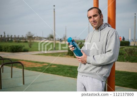 Determined young athlete holding water bottle, resting after outdoor workout on an urban sportsground, looking at camera 112137800