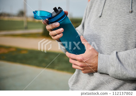 Selective focus on the water bottle in the hands of an active male athlete resting after workout or running outdoor Selective focus on the water bottle in the hands of an active male athlete resting after workout or running outdoor 112137802