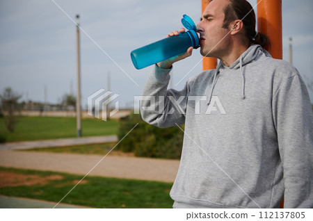 A sportsman drinking water from the bottle, hydrating and resting after heavy workout on the urban sportsground A sportsman drinking water from the bottle, hydrating and resting after heavy workout on the urban sportsground 112137805