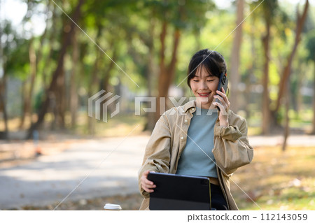 Smiling young woman freelancer sitting on park bench with digital tablet and talking on mobile phone Smiling young woman freelancer sitting on park bench with digital tablet and talking on mobile phone 112143059