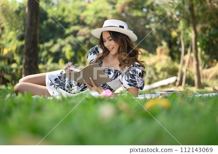 Serene young lady enjoying new novel, sitting on green grass in the park on sunny day 112143069