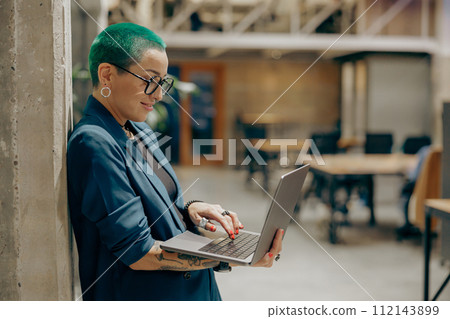 Pretty tattooed female manager with green short hair is use laptop standing in modern coworking  112143899