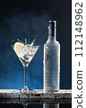 Vodka Cocktail glass splash and ice-cold bottle on blue degraded background with copy space. Vertical format. 112148962