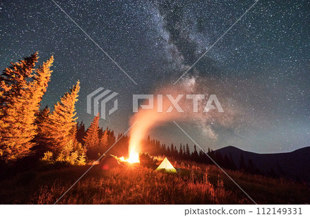Night camping under sky full of stars and Milky way in the mountains. Starry sky over illuminated tourist tents on hills near forest. Warm light from campfire at dark night. Night camping under sky full of stars and Milky way in the mountains. Starry sky over illuminated tourist tents on hills near forest. Warm light from campfire at dark night. 112149331
