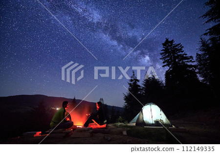 Night-time sojourn in mountains, beneath starry sky. Man and woman, intrepid wanderers, unwind by flickering flames of campfire, alongside their tent pitched near the serene forest, under Milky way. Night-time sojourn in mountains, beneath starry sky. Man and woman, intrepid wanderers, unwind by flickering flames of campfire, alongside their tent pitched near the serene forest, under Milky way. 112149335