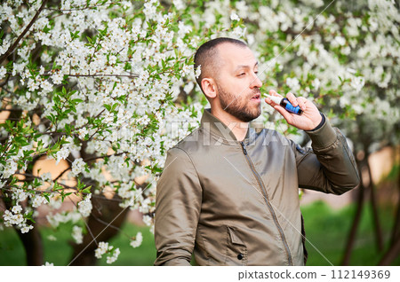Man allergic using medical nasal drops, suffering from seasonal allergy at spring in blossoming garden. Handsome man treating runny nose in front of blooming tree outdoors. Spring allergy concept. Man allergic using medical nasal drops, suffering from seasonal allergy at spring in blossoming garden. Handsome man treating runny nose in front of blooming tree outdoors. Spring allergy concept. 112149369