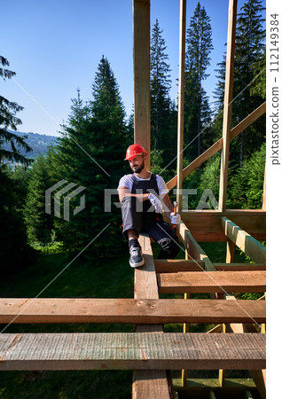 Carpenter constructing wooden frame two-story house near the forest. Bearded man holding hammer, dressed in protective helmet and overalls. Concept of ecological modern construction. Carpenter constructing wooden frame two-story house near the forest. Bearded man holding hammer, dressed in protective helmet and overalls. Concept of ecological modern construction. 112149384