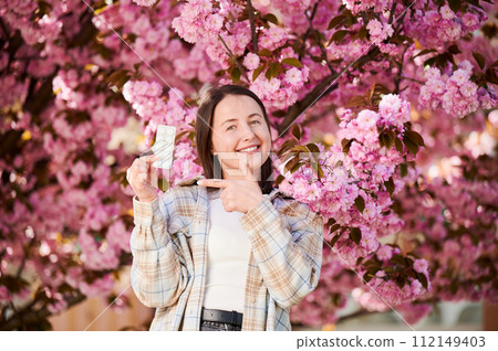 Woman allergic suffering from seasonal allergy at spring. Young happy woman showing pack of pills, posing in blossoming garden at springtime. Antihistamine medication concept 112149403