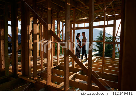 Man and woman inspecting their future wooden frame house nestled in the mountains near forest. Youthful couple at construction site in early morning. Concept of contemporary ecological construction. 112149411