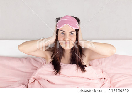 Woman cannot sleep, she covers her ears with her hands, the neighbors are noisy, making repairs. Pink sleep mask, pink bed linen. Woman cannot sleep, she covers her ears with her hands, the neighbors are noisy, making repairs. Pink sleep mask, pink bed linen. 112150290