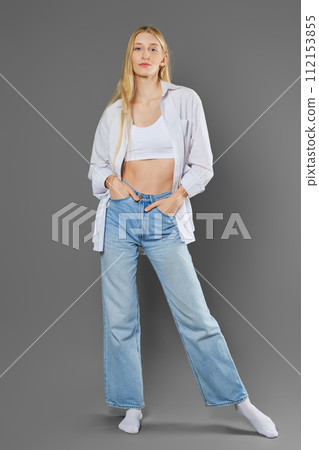 A tall woman in a tank top, unbuttoned shirt, jeans and socks posing on a grey background 112153855