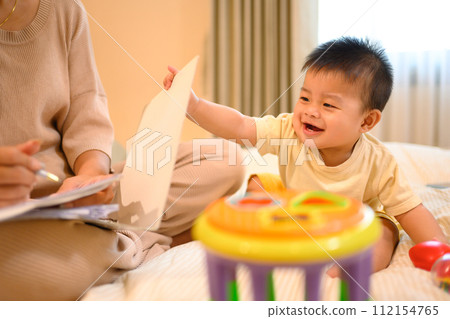 Cheerful little baby boy sitting on the bed near busy mother working on laptop Cheerful little baby boy sitting on the bed near busy mother working on laptop 112154765