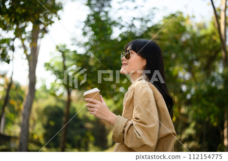 Cheerful dreamy woman in sunglasses holding paper cup relaxing in the forest park Cheerful dreamy woman in sunglasses holding paper cup relaxing in the forest park 112154775