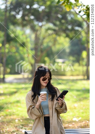 Portrait of young stylish woman in sunglasses drinking coffee and using mobile phone in the park 112154808