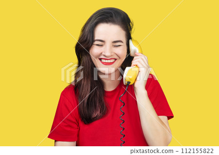 A beautiful brunette woman with red lipstick speaks on an old yellow telephone with a cord. She is smiling and happy. on a yellow background A beautiful brunette woman with red lipstick speaks on an old yellow telephone with a cord. She is smiling and happy. on a yellow background 112155822