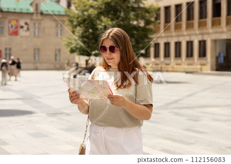 Happy tourist girl with map travels in Europe. Vacation concept by exploring interesting places to travel. Redhead Woman Searching locations at summer sunny day 112156083
