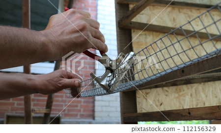 male hands with pincers bend the mesh material into the structure of a bird cage during manufacturing, making a cage for quails from a metal lattice and a wooden frame male hands with pincers bend the mesh material into the structure of a bird cage during manufacturing, making a cage for quails from a metal lattice and a wooden frame 112156329
