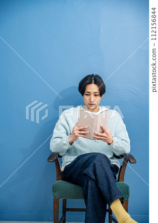 A young man sitting on a chair and reading a book against a blue background 112156844