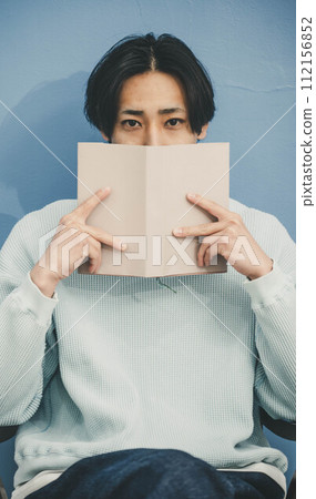 A young man sitting on a chair and reading a book against a blue background A young man sitting on a chair and reading a book against a blue background 112156852