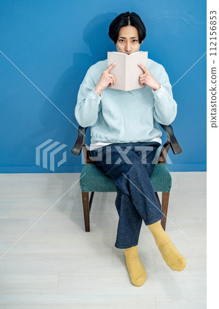 A young man sitting on a chair and reading a book against a blue background 112156853