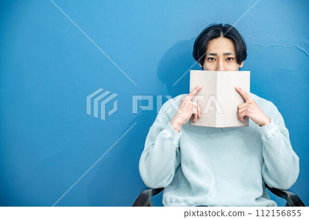 A young man sitting on a chair and reading a book against a blue background A young man sitting on a chair and reading a book against a blue background 112156855