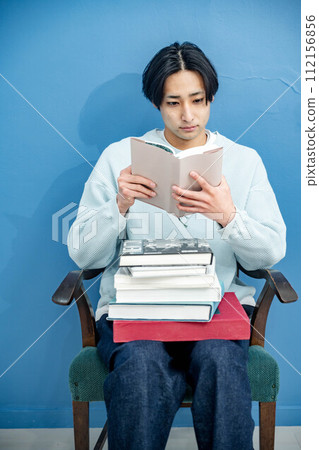 A young man sitting on a chair and reading a book against a blue background 112156856