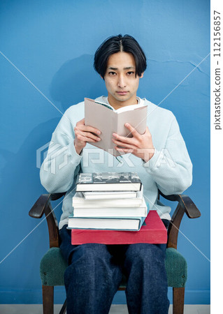 A young man sitting on a chair and reading a book against a blue background 112156857