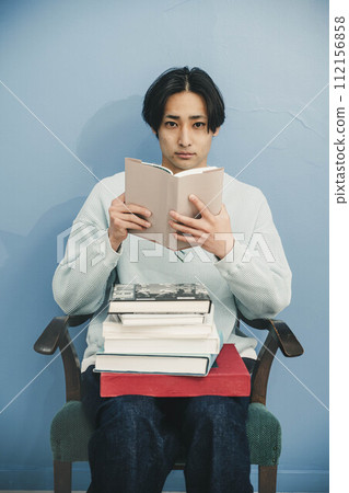 一個年輕人坐在椅子上,在藍色背景下讀書 一個年輕人坐在椅子上,在藍色背景下讀書 112156858