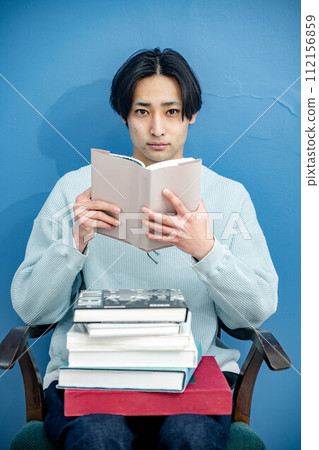 A young man sitting on a chair and reading a book against a blue background A young man sitting on a chair and reading a book against a blue background 112156859