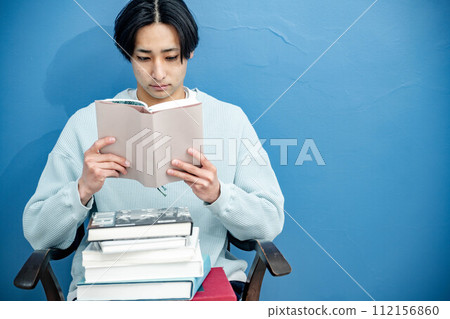 A young man sitting on a chair and reading a book against a blue background 112156860