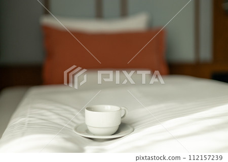 Coffee or tea cup on the bed. Hotel room or bedroom Interior, morning time. Concept of easy breakfast. One white ceramic cup. 112157239