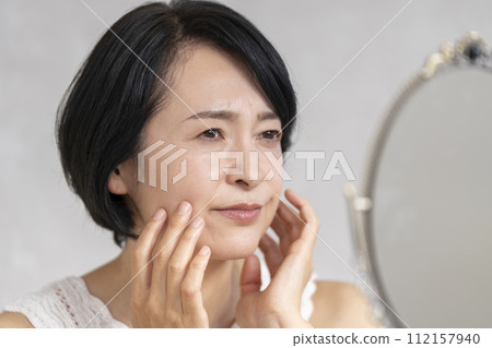 A woman in her early 50s puts her hand on her face while looking in the mirror. Troubles and skin problems. Beauty image. A woman in her early 50s puts her hand on her face while looking in the mirror. Troubles and skin problems. Beauty image. 112157940