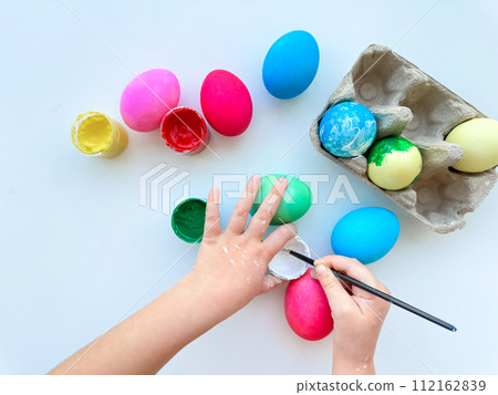 Childrens hands paint Easter eggs with brush surrounded by colorful eggs and jars of paints on white table, creative holiday activity. Childrens hands paint Easter eggs with brush surrounded by colorful eggs and jars of paints on white table, creative holiday activity. 112162839