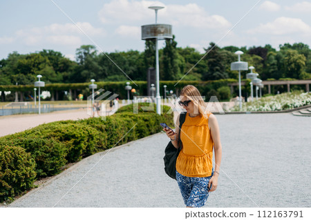 Happy cheerful young blond woman with backpack walking on city street checks her smartphone. Portrait of beautiful 30s girl using smartphone outdoors. Urban lifestyle concept. Summer time Happy cheerful young blond woman with backpack walking on city street checks her smartphone. Portrait of beautiful 30s girl using smartphone outdoors. Urban lifestyle concept. Summer time 112163791