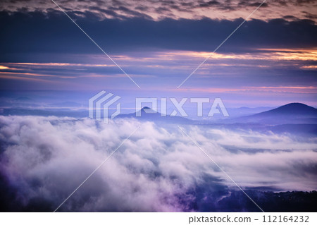 Tranquil sunrise with a lot of fog under orange-purple colorful sky, volcanic mountains in Central Bohemian Uplands (Czech: Ceske Stredohori). Impressive foggy morning landscape panorama. Tranquil sunrise with a lot of fog under orange-purple colorful sky, volcanic mountains in Central Bohemian Uplands (Czech: Ceske Stredohori). Impressive foggy morning landscape panorama. 112164232