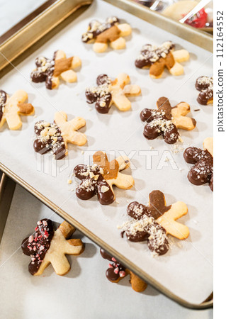 Creating cutout sugar cookies, partially dipped in chocolate and topped with hazelnut pieces, placed on parchment paper. Creating cutout sugar cookies, partially dipped in chocolate and topped with hazelnut pieces, placed on parchment paper. 112164552