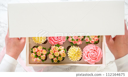 Flat lay. In a woman's hands, a white paper box filled with gourmet cupcakes, each adorned with vibrant roses and tulips crafted from buttercream frosting, is gently being closed. 112164675