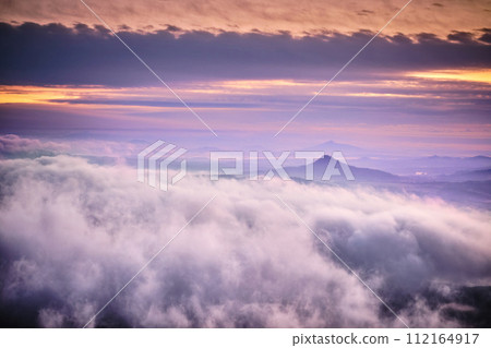 Tranquil sunrise with a lot of fog under orange-purple colorful sky, volcanic mountains in Central Bohemian Uplands (Czech: Ceske Stredohori). Impressive foggy morning landscape panorama. Tranquil sunrise with a lot of fog under orange-purple colorful sky, volcanic mountains in Central Bohemian Uplands (Czech: Ceske Stredohori). Impressive foggy morning landscape panorama. 112164917