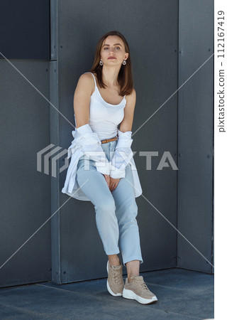 Stylish young woman at light casual outfit standing near the gray wall of the modern building. 112167419