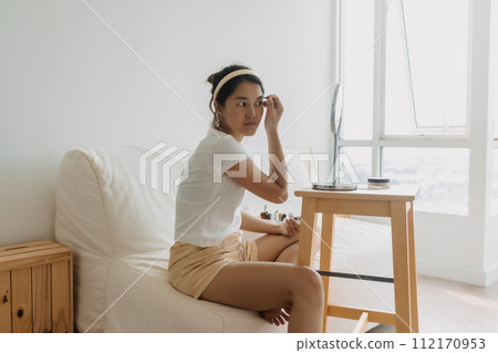 Asian Thai woman using pencil and drawing eyebrow, sitting on white couch in front of the mirror in room apartment, facial makeup and cosmetic skin concept. Asian Thai woman using pencil and drawing eyebrow, sitting on white couch in front of the mirror in room apartment, facial makeup and cosmetic skin concept. 112170953