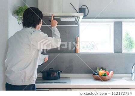 A male businessman in work clothes inspects the range hood and ventilation fan in the kitchen. 112172754
