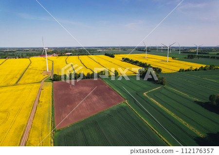 Aerial view Wind turbine on grassy yellow farm canola field against cloudy blue sky in rural area. Offshore windmill park with clouds in farmland Poland Europe. Wind power plant generating electricity 112176357