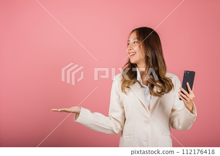 Happy Asian portrait beautiful cute young woman excited holding mobile phone blank screen presenting product with palm of hand at empty, studio shot isolated on pink background, female look to space 112176418