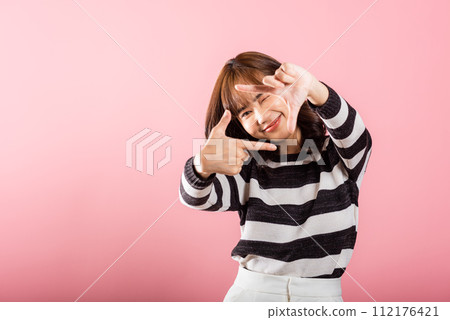 A cheerful Asian photographer frames her vision with a finger, expressing the concept of photography. Studio shot on pink background, highlighting inspiration and creativity. 112176421
